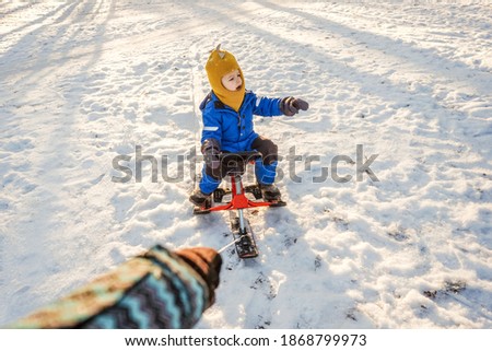 Womans hand rolls a small child boy on a snowmobile through the snow in the park. Sun light and trees shadows on background.