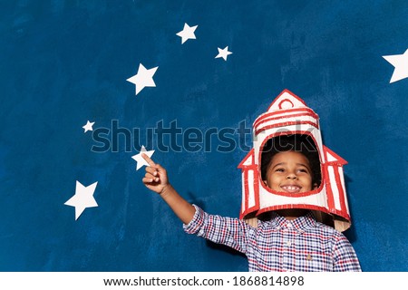 Rocket boy, afro-american kid playing astronaut pointing aside upwards on blue background with white hand-made stars. Joyful child, future cosmonaut with spaceship made out of cardboard.