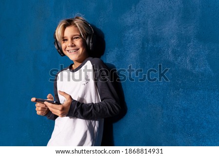 Laughing american teenage boy in headphones using phone app on blue background with copy space. Smiling schoolboy listening lecture, music or audiobook using headset. Education and technology concept.