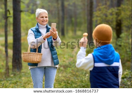 picking season, leisure and people concept - happy smiling grandmother with smartphone photographing grandson with mushrooms in basket in forest