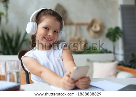 Smart little girl in headphones do homework online class on smartphone, small child in earphones study on Internet, have web conference or lesson during quarantine.