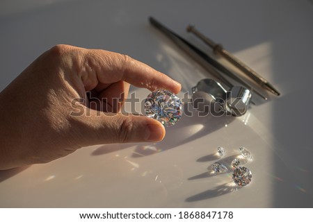 Close-up of the hand of jeweller gemologist with large size round cut diamond on desktop with magnifier and tweezers. Buyer diamond expert checking color of diamond in natural light. Royalty-Free Stock Photo #1868847178