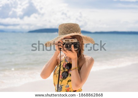 Short-haired Asian woman in a yellow floral dress and straw hat standing on the beach using camera take a photo in front of her with a blurry sea, mountain and cloudy sky background on a sunny day.