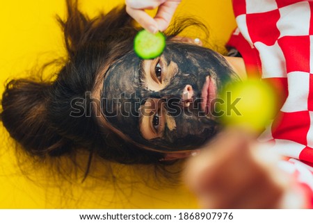 Girl with black face mask and cucumbers on her eyes. Relaxing, spa, charcoal mask eliminates black spots. Smooth yellow background. Anti-stress, relaxing
