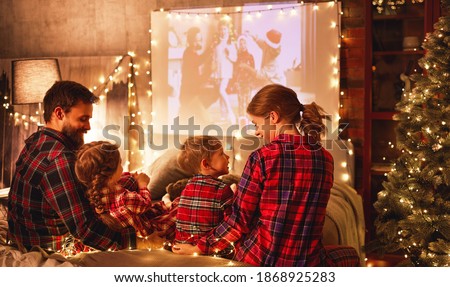 happy family in checkered pajamas: mother father and children watching projector, film, movies with popcorn in christmas holiday evening   at home 
