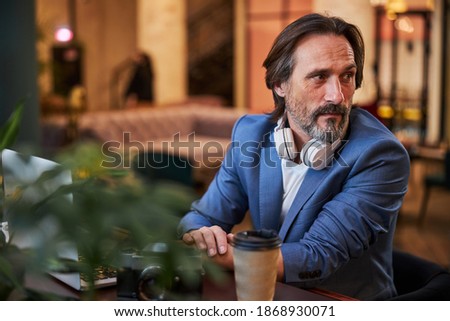 Concerned brunette man in elegant blazer looking to the side while sitting at a comfortable table with his laptop