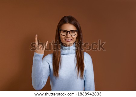 Excited punk young caucasian woman makes rock gesture, looks cool, expresses excitement and happiness, has appealing appearance, wears glasses and light blue sweater, isolated on brown background