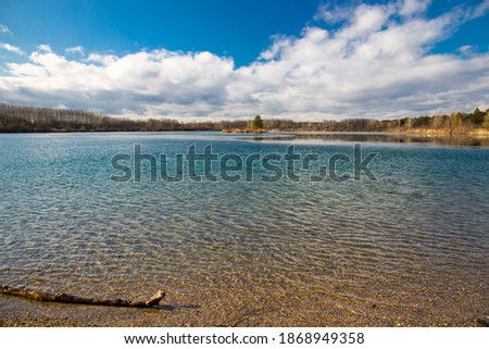 Blue cold waters of Cunovo lake with the blue cloudy sky and dry autumn trees around the lake, and tree branch on the left side of the picture
