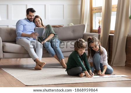 Happy little brother and sister kids children sit on floor in new home drawing painting together. Young Caucasian parents relax on couch using tablet pad. Family rest in own house on weekend.