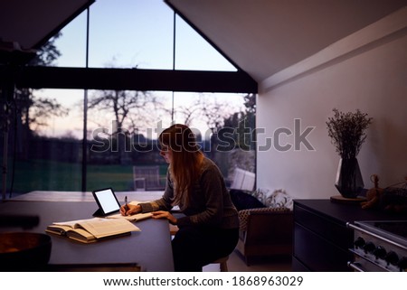 Evening Shot Of Woman In Kitchen Working Or Studying From Home Using Digital Tablet Royalty-Free Stock Photo #1868963029