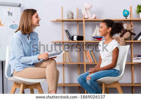 Smiling african american girl looking at psychologist with digital tablet during consultation with blurred office on background