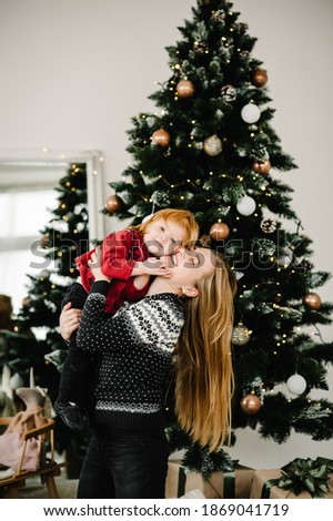 Merry Christmas and Happy Holidays. Cheerful mom hugging cute baby daughter girl near Christmas tree. Mother and little child having fun and playing together at home.
