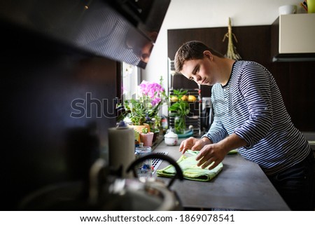 Down syndrome adult man standing indoors in kitchen at home, helping. Royalty-Free Stock Photo #1869078541