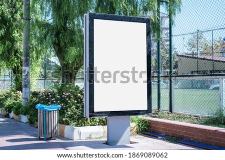 Big empty billboard in black frame with empty white mockup space on sidewalk of city street in summer day