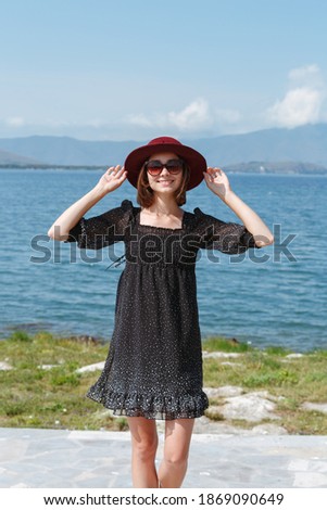 Charming woman wearing short dress with polka dot pattern standing on shore of sea and touching hat while smiling and enjoying summer holiday at seaside 