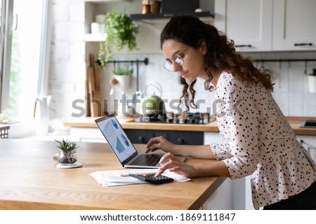 Serious young woman wearing glasses calculating finances, household expenses, confident businesswoman working with project statistics, using laptop and calculator, standing in kitchen at home Royalty-Free Stock Photo #1869111847