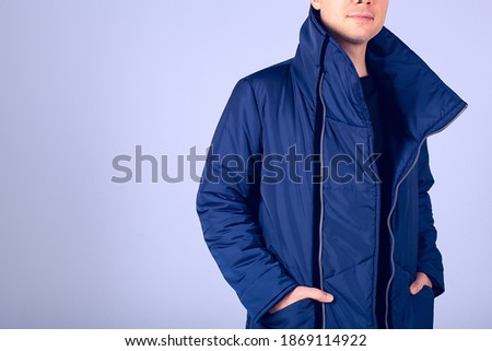 The guy model on a gray background is dressed in a blue down jacket. Hands in the pockets of a warm jacket. A guy with a beard and warmly dressed. Stylish warm clothes