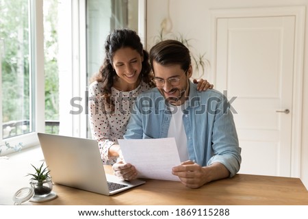 Happy wife and husband wearing glasses reading good news in letter, sitting at table with laptop, checking financial documents, received mortgage or loan approve notification, planning budget Royalty-Free Stock Photo #1869115288