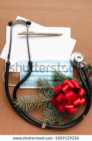 blue medical mask mockup, medical phonendoscope, christmas tree branch, blank sheet decorated with red bow on isolated background with place for text