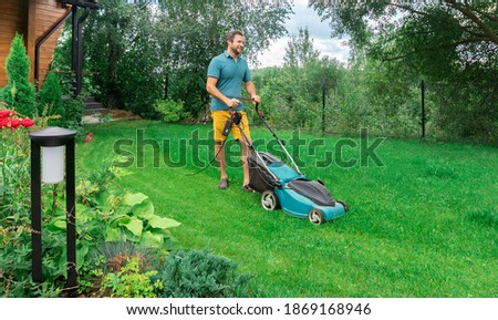 A man in a good mood mows the grass with an electric lawn mower in the garden. The hardworking owner takes care of the lawn on a sunny summer day. Royalty-Free Stock Photo #1869168946