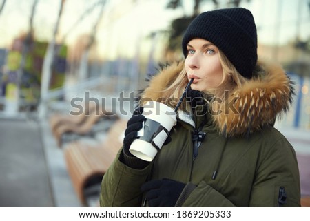 Attractive young hipster woman with a natural make-up in a knitted warm winter hat in a sweatshirt in a stylish jacket with fur holds a cup of coffee in her hand. Cute girl.