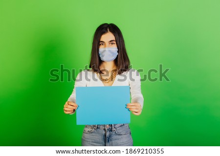 Serious woman in medical mask showing empty blue blank paper in her hands. Woman standing on green background. Empty space for advertisement. Coronavirus.