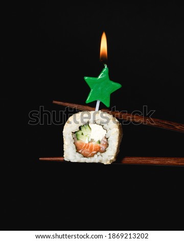 New Year sushi with candle on black background. Philadelphia maki roll piece between chopsticks. Green star shaped burning candle. Christmas celebration party. Creative japanese food. Faded, close up