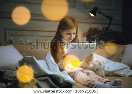 Dreams. Happy caucasian little girl during video call with laptop and home devices, looks dreamful and happy. Talking to Santa before New Year's eve, her family, watching cartoons, typing text. Bokeh