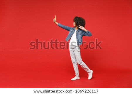 Full length side view little african american kid girl 12-13 years old in denim jacket doing selfie shot on mobile phone showing victory sign isolated on red background. Childhood lifestyle concept