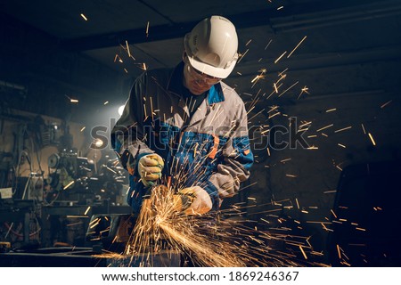 Locksmith in special clothes and goggles works in production. Metal processing with angle grinder. Sparks in metalworking. Royalty-Free Stock Photo #1869246367