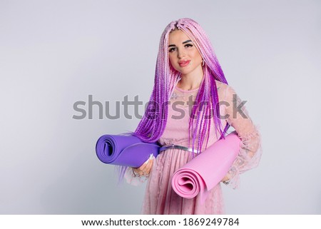 Cute girl in elegant dress with pink hair and two yoga mats is smiling against a white background with a side space.