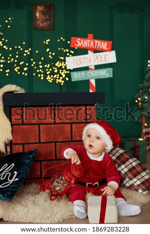 Little boy in a red Santa Claus suit is sitting and playing near a decorative chimney.