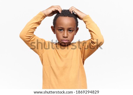 Studio image of sad upset dark skinned little boy posing isolated keeping hands on his curly short hair. Unhappy displeased Afro American child scratching head, feeling uncomfortable, worrying
