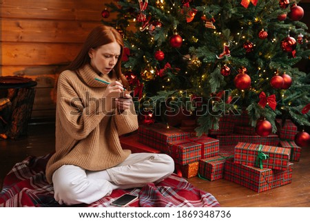 Dreamy redhead young woman writing Christmas letter to Santa Claus sitting on floor at home on background of xmas tree at cozy living room with festive interior. Concept of home festive atmosphere.