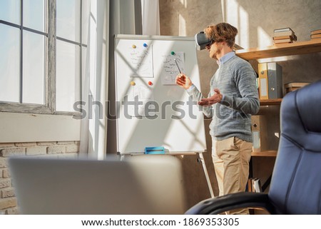 Businessman wearing virtual reality glasses standing near dashboard while working at home and making online presentation for people. Stock photo