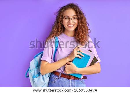 happy school girl with bag and books posing at camera isolated over purple background, cheerful female with curly hair smile, happy to go school. education concept Royalty-Free Stock Photo #1869353494