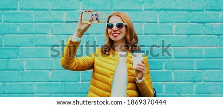 Portrait of happy smiling young woman taking selfie picture by smartphone wearing a yellow jacket over blue background