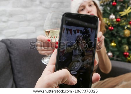 Cheerfull young woman in Santa hat posing in front of phone camera with a glass of wine. Christmas or New Year Eve celebration at home.