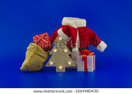 Red Santa Claus suit near wooden Christmas tree with Santa hat and gift boxes on a festive blue background. New Year and Christmas gift season concept