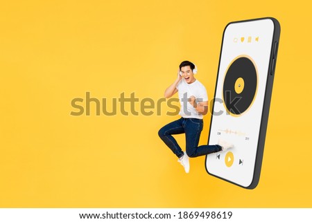 Happy young Asian man wearing headphones listening to music from mobile phone application and jumping on yellow studio background with copy space