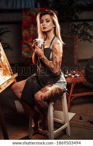 Blond haired slim female painter with apron and coffee cup poses in dark workshop sitting on chair around artworks.