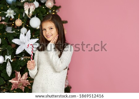 adorable little child girl holding lollipop candy cane on the pink background of christmas tree. copy space