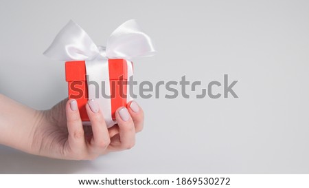 The hand of a woman with a pink manicure holds a red gift box with a white ribbon on a gray background: space for text