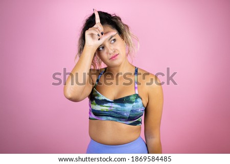 Young beautiful woman wearing sportswear over isolated pink background making fun of people with fingers on forehead doing loser gesture mocking and insulting.