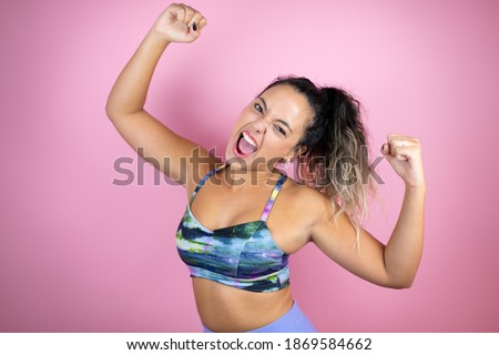Young beautiful woman wearing sportswear over isolated pink background very happy and excited making winner gesture with raised arms, smiling and screaming for success.