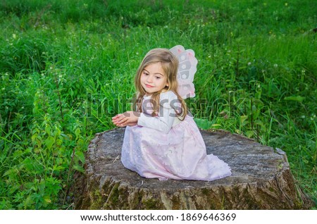 Little cute girls with butterfly wings on a large tree stump among the tall green grass. Selective focus. Blurred background.