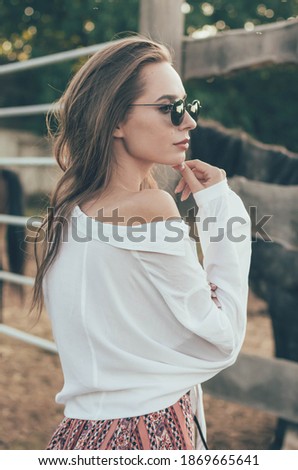 
a woman of model appearance with long hair dressed in a shirt, a long skirt stands near the fence in sun glasses on the background of an equestrian farm near the horses at sunset