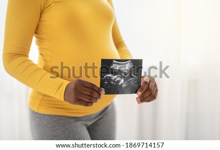 Unrecognizable pregnant african american woman holding ultrasound image of her unborn child, standing by window at home, copy space. Cropped of black lady with big tummy showing sonogram