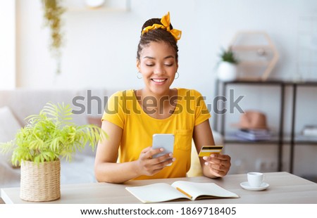 Online Order And Ecommerce Concept. Smiling African American Woman Using Smartphone And Holding Credit Card, Sitting At Desk With Open Notebook In Living Room At Home. Internet Shop Offers And Sales Royalty-Free Stock Photo #1869718405