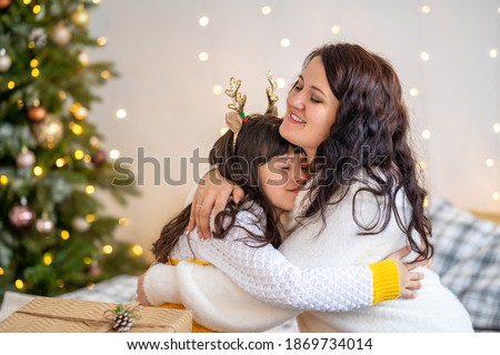Merry Christmas and Happy Holidays! Happy mom and daughter hugging, and hold with christmas gift box on background Christmas tree decorated with a garland. Copy space